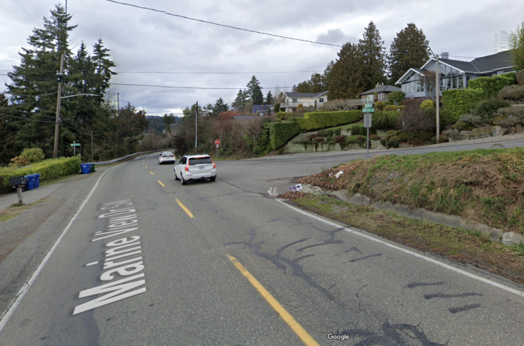 Street view photo looking north on Marine View Drive as it approaches 46th Ave SW. 46th is a significant downhill toward the intersection, and Marine View is a slight downhill. 46th is the only street with a stop sign, and it intersects at an obtuse angle.